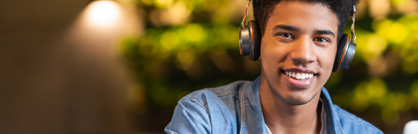 A student wearing headphones smiles at the camera from behind his laptop.