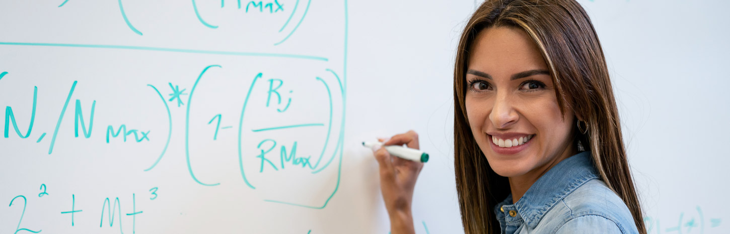 A woman writes mathematical formulas on a whiteboard while looking over her shoulder and smiling at the camera.