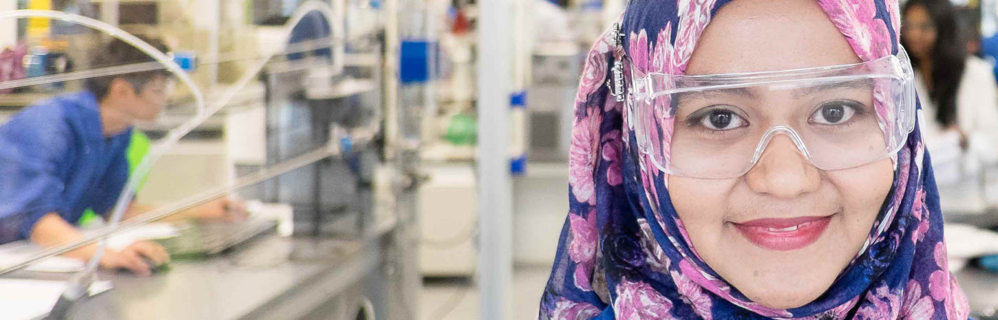 A young woman in a hijab stands in front of scientific equipment and smiles at the camera.